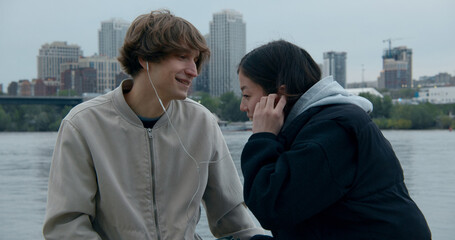 Young couple sharing earbuds while listening to music by the river