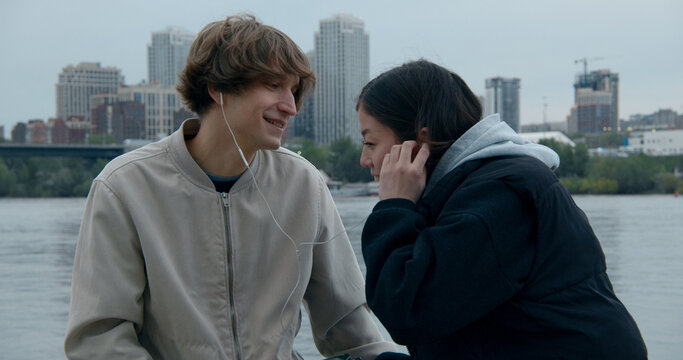 Young couple sharing earbuds while listening to music by the river