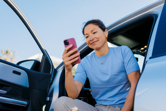 Smiling woman using smartphone while sitting in car with open door