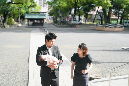 In June 2025, a young Japanese mother and father in their early twenties walk past the shrine's main hall, holding their one-month-old baby and honoring the traditional omiyamairi ceremony in Aichi.