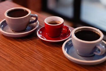 Three cups on a wooden table: two larger, light blue cups filled with black coffee on matching saucers, and a smaller, vibrant red cup filled with espresso on a red saucer