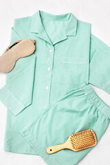 Flat lay with green Pajama, beige sleep eye mask, hair brush on white sheet. Top view trend mint...