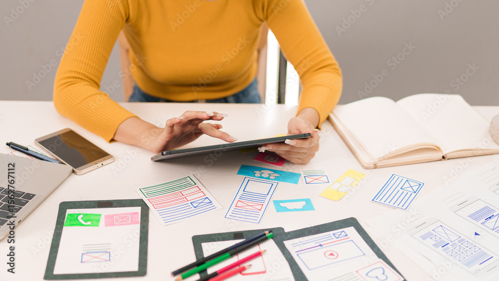 Wall mural adult asian woman ui designer working on tablet with user interface wireframes and sketches for mobi