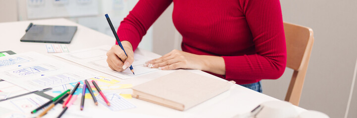 Asian woman UI UX designer sketches mobile app interfaces user experience planning website layout design on paper prototype at desk for startup