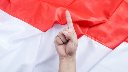 A hand making a gesture with the index finger pointing upward symbolizes number one or unity in front of the red and white Indonesian flag to celebrate Indonesia independence day