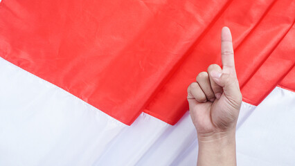 A hand making a gesture with the index finger pointing upward symbolizes number one or unity in front of the red and white Indonesian flag to celebrate Indonesia independence day