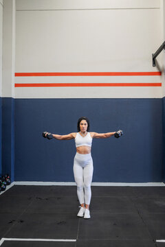 Young Latina doing lateral raises with dumbbells at the gym