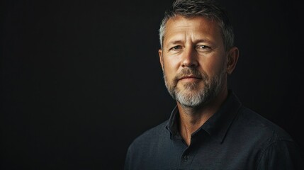 Fototapeta premium A man with gray hair and a beard, wearing a dark shirt, standing against a black background.