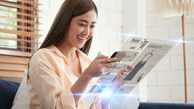 Happy Asian woman shopping online with smartphone. Futuristic holographic interface showing clothing items and digital icons, concept of smart fashion e-commerce and digital retail lifestyle.