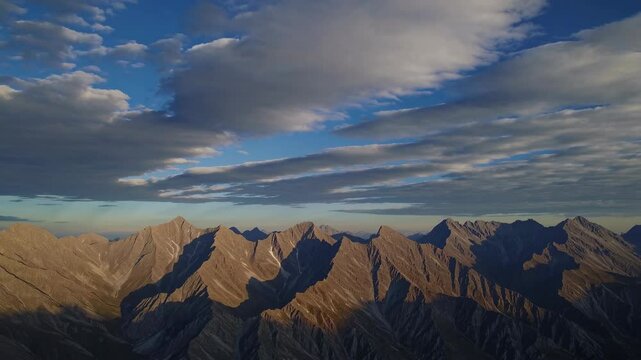Majestic mountain peaks loom under a sky filled with dramatic clouds at sunset. The warm hues reflect off the rugged terrain, creating a stunning natural panorama