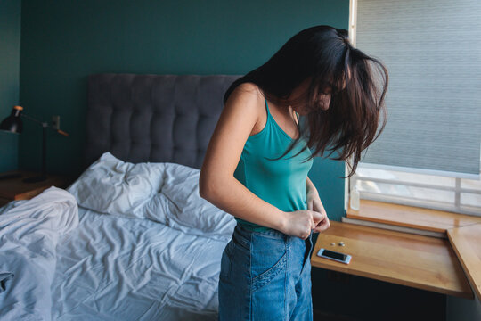 Young brunette woman getting ready buttoning jeans in bedroom 