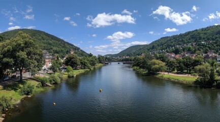 Obraz premium Panoramic view of a river flowing between verdant hills, lined with quaint buildings and lush trees under a bright, partly cloudy sky