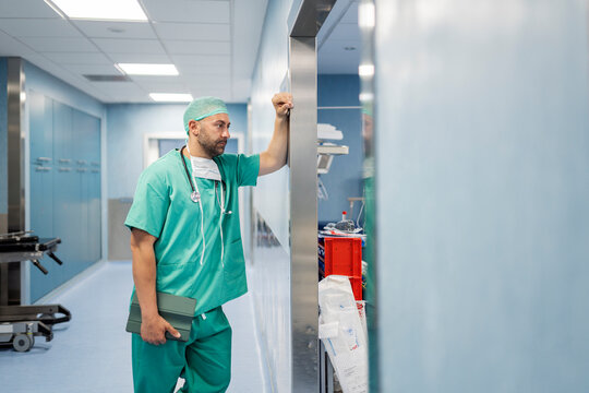 Thoughtful surgeon waiting in hospital corridor holding tablet - Powered by Adobe