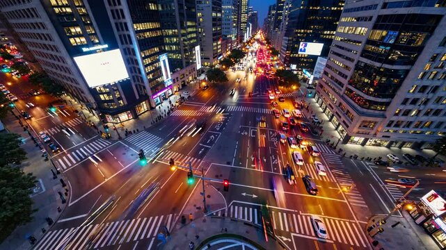 An overhead view captures a bustling urban intersection at night, with city lights illuminating the streets and traffic flowing among tall buildings. The atmosphere exudes energy