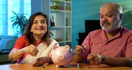 Indian senior couple promoting saving money investment habits for future security, mature husband wife placing coins in piggy bank at home, planning retirement, budget, financial awareness concept