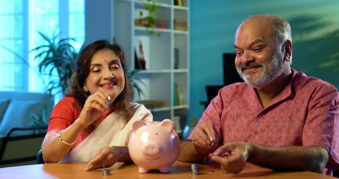 Indian senior couple promoting saving money investment habits for future security, mature husband wife placing coins in piggy bank at home, planning retirement, budget, financial awareness concept - Powered by Adobe
