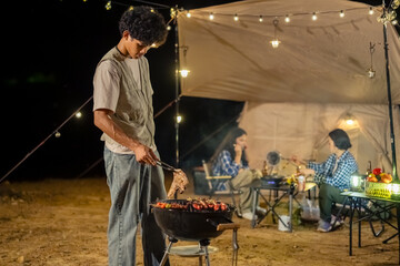 A man is cooking food on a grill outside. There are three people around the grill, one of whom is holding a fork