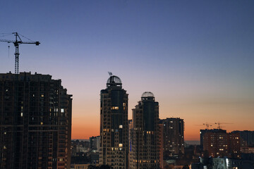 Fototapeta premium Urban skyline at dusk featuring modern high-rise buildings with construction cranes silhouetted against a vibrant sunset, showcasing architectural development and city life