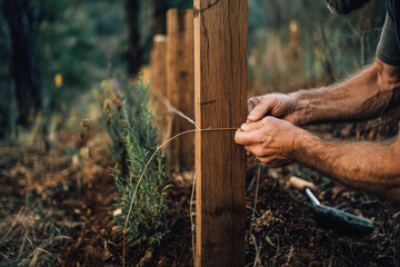 A close-up of a man's hands tying wire around a wooden fence post in a natural setting. The scene emphasizes off-grid living and sustainable practices.