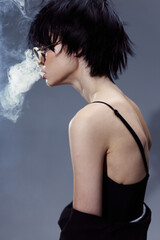 young woman in black top wearing glasses exhales smoke in moody studio lighting with dark gray...