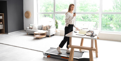 Young businesswoman with coffee cup training on treadmill at table in office