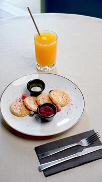 Traditional Eastern European oladyi pancakes presenting golden surface, accompanied by tangy sour cream, vibrant berry sauce, and fresh seasonal berries beside orange juice