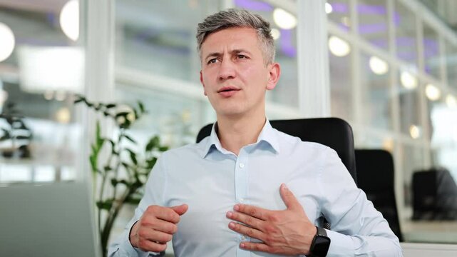 Professional businessman feeling intense chest pain and discomfort, touching his chest and unbuttoning his shirt in a modern office environment. He appears stressed.