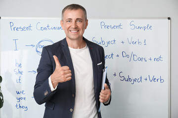 Male teacher with clipboard showing thumb-up gesture near flipboard in classroom