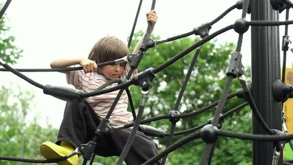 A child boy is climbing up an alpine grid in a park on a playground on a hot summer day. Kids playground in a public park, entertainment and recreation for children, mountaineering training. - Powered by Adobe