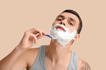 Handsome young man with razor and shaving foam on his face against beige background