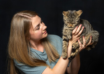 Portrait of cheerful woman holding small striped cat close, eyes full of affection. Minimalist dark...