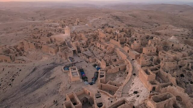 A desert city with ancient ruins and structures, including buildings and rocky outcrops. The sun is setting over the landscape, casting warm light on the area. Location: Tunisia, Gabes , Zraoua.