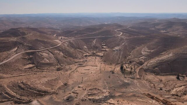 A road that has been left to deteriorate over time. It is surrounded by desert mountains with barren trees and dirt landscapes. The sky can be seen above the area, giving it an open and vast feeling.