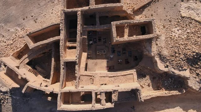 A ruined stone building with many steps leading to a large chamber. The area is filled with stones and has an open area. There are people around the site, likely exploring or visiting this historical