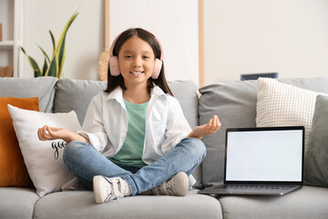 Little Asian girl in headphones with laptop meditating on sofa at home