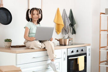 Little Asian girl with headphones using laptop in kitchen