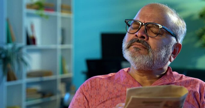 Indian man solving crossword puzzle with pencil while sitting at table, elderly Asian male reading newspaper, focused relaxed, enjoying peaceful morning brain exercise as part of daily routine