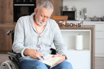 Senior man suffering from Parkinson syndrome in wheelchair drawing on notebook at home