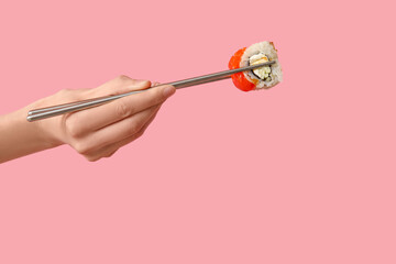 Female hand holding chopsticks with tasty sushi roll on pink background