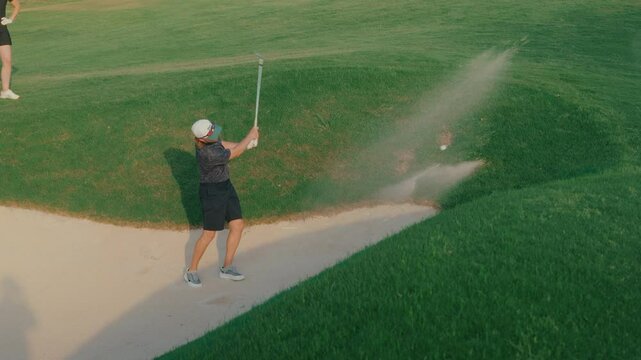 Handheld shot of a male golfer hitting from a greenside bunker in slow motion as his playing partner watches from nearby. Sand flies as the ball lifts, capturing a focused short game moment.