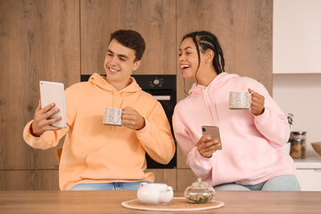 Young couple with cups of coffee using tablet computer and mobile phone in kitchen