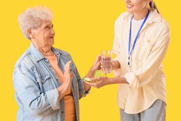 Senior woman refusing to take pills from nurse on yellow background