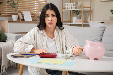 Tired young woman with piggy bank and calculator doing taxes at home. Stress concept