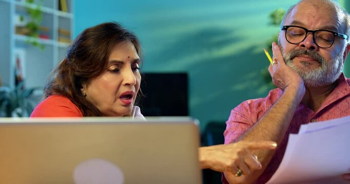 Indian couple managing home finance using calculator and documents, senior Asian husband wife sitting together at table reading bills, planning retirement pension, doing paperwork and monthly budget