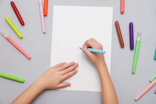 Child's hands drawing with felt-tip pens and sheet of paper on grey background. Top view