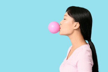 Pretty young Asian woman blowing pink bubble gum on blue background