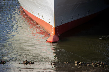 Ship's bow grounded in shallow water at low tide, showing the red hull and muddy shoreline....