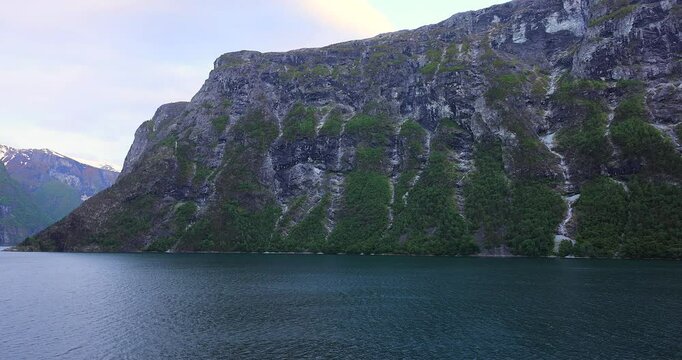 Majestic green cliffs and deep blue fjord waters under cloudy skies, filmed from a cruise ship sailing through the breathtaking landscapes of Norway fjords.