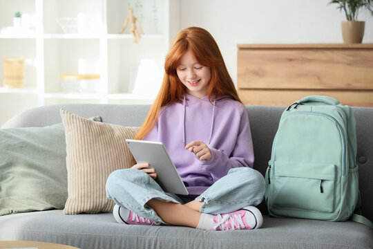 Cute ginger girl with backpack and modern tablet computer sitting on sofa at home