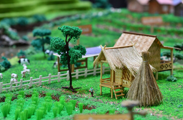 Miniature rural farming scene featuring traditional thatched-roof hut, rice field, livestock and...